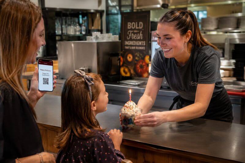 Kids birthday freebie celebration at a restaurant with free dessert reward - MoneyPantry.com Child receiving a free birthday treat at a restaurant while a parent shows a birthday reward on a smartphone